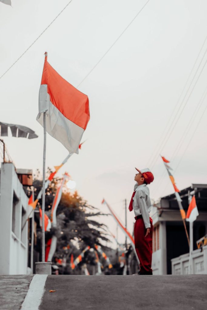 a boy standing in uniform in front of flag of indonesia hanging on a pole